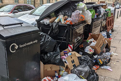 Excess rubbish piled around large bins.